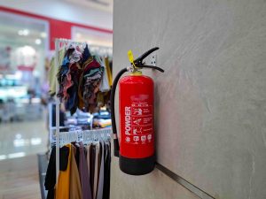 Extintor de incêndio fixado na parede de loja de roupas, ao lado de peças de vestuário, ilustrando AVCB para comércios