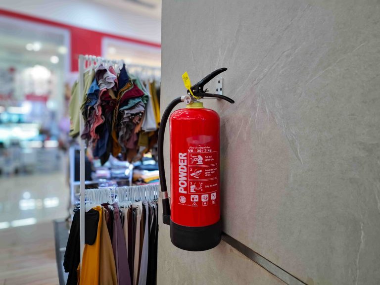 Extintor de incêndio fixado na parede de loja de roupas, ao lado de peças de vestuário, ilustrando AVCB para comércios