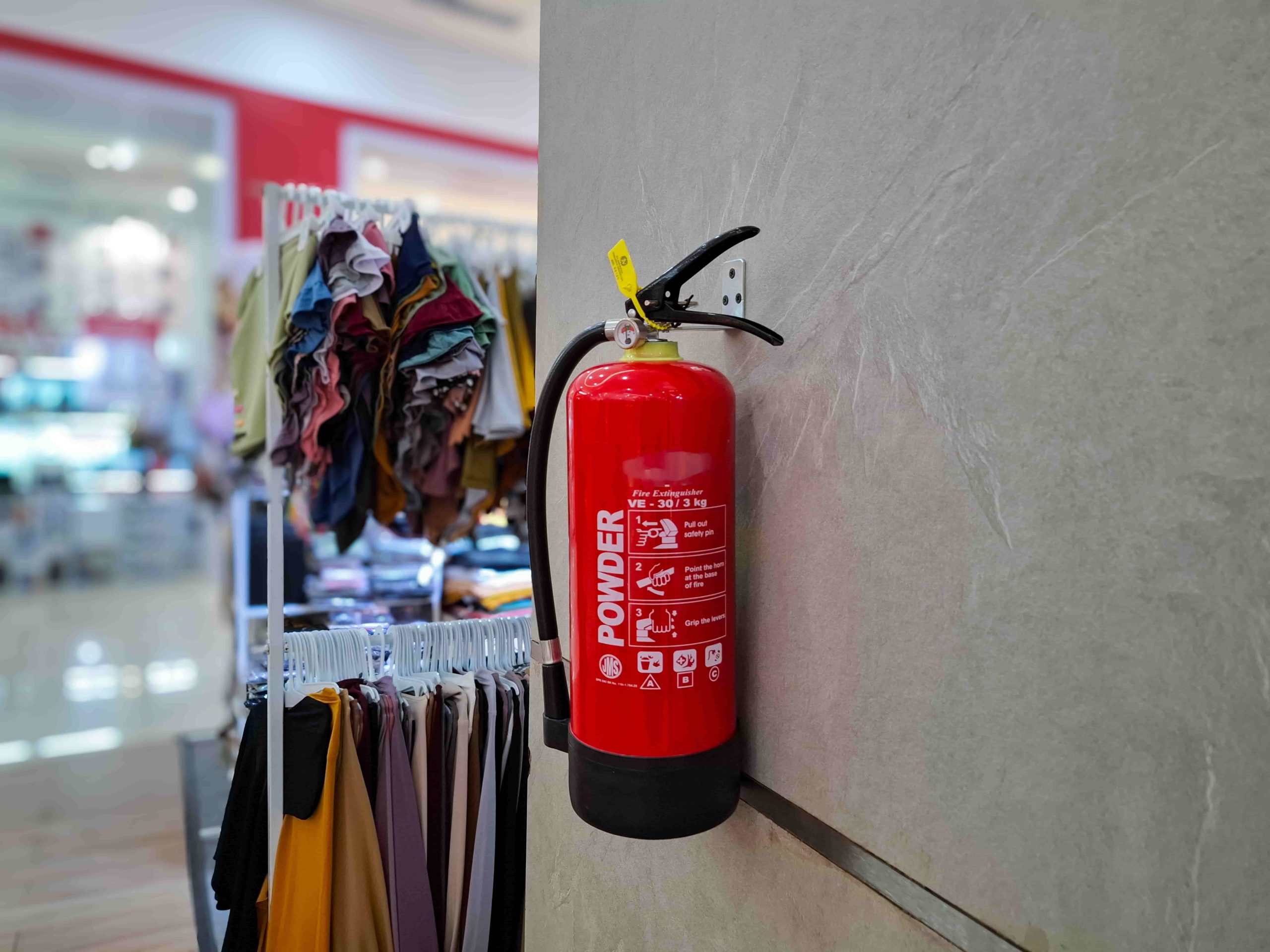 Extintor de incêndio fixado na parede de loja de roupas, ao lado de peças de vestuário, ilustrando AVCB para comércios